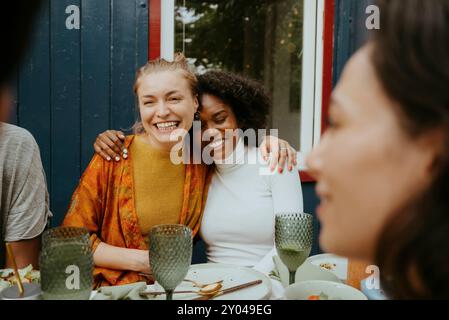 Happy multiracial female friends sitting with arms around while talking to friends at dinner party Stock Photo