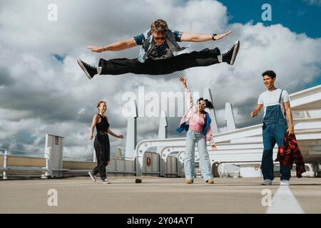 Young man doing split in mid-air while male and female friends standing in parking lot under cloudy sky Stock Photo