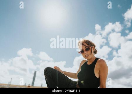 Portrait of young woman in sunglasses and black casuals posing under cloudy sky at sunny day Stock Photo