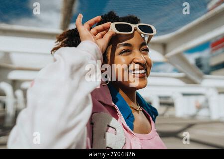 Happy fashionable curly hair woman in letterman jacket holding sunglasses at parking lot Stock Photo