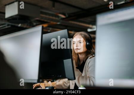 Focused female entrepreneur with wireless headphones doing coding on computer in office Stock Photo