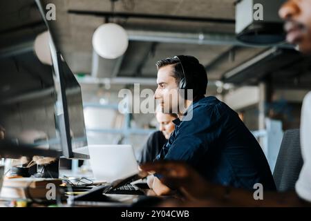 Young programmer with wireless headphones sitting and looking at computer in office Stock Photo