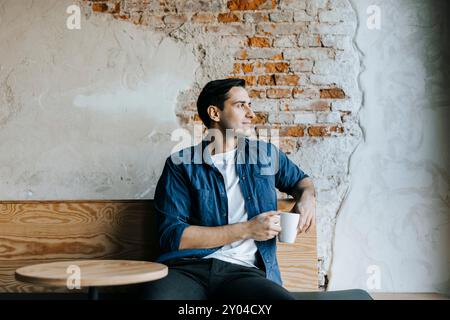 Young tech entrepreneur with coffee cup looking away while sitting against brick wall in office Stock Photo