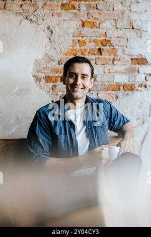 Portrait of smiling young programmer sitting with coffee cup against brick wall in office Stock Photo