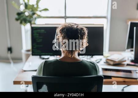 Rear view of female programmer with curly hair wearing headphones working in office Stock Photo