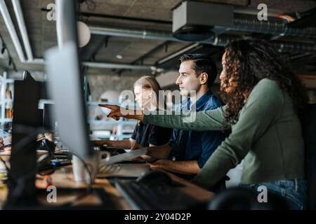 Female coder pointing at device screen while sitting with colleagues in office Stock Photo