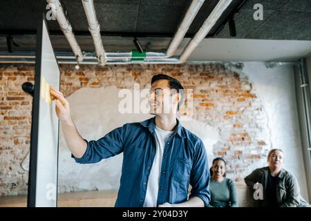 Smiling male tech professional sticking adhesive notes on whiteboard in office Stock Photo
