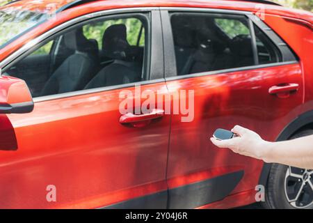 A close-up of a hand holding a car key fob next to a red vehicle, concept of remote car access and control. Everyday moments associated with car owner Stock Photo