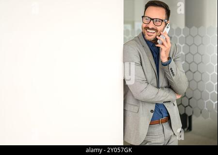 Successful male manager talking over smartphone and looking away while leaning on white wall in office Stock Photo