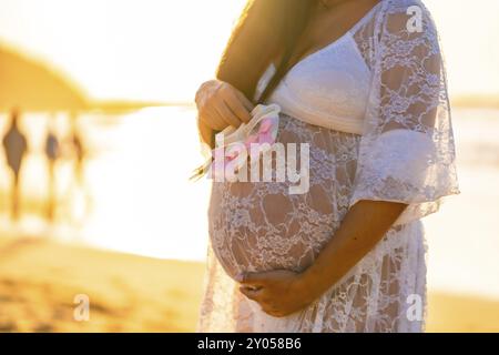 A woman is wearing a white lace dress and holding a flower in her hand. The scene is set on a beach, with the sun shining brightly in the background Stock Photo