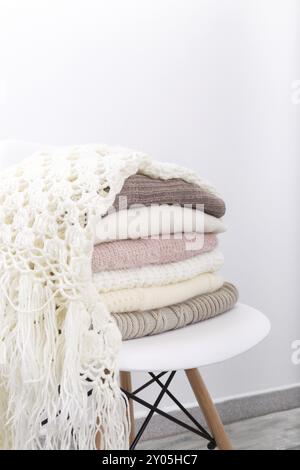Stack of cozy knitted sweaters or blankets pastel trending colors on white modern chair against the wall Stock Photo