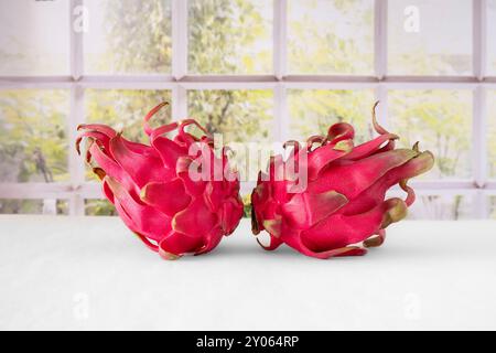 Close-up of whole and cut red dragon fruit on a table in front of a window with plants behind it Stock Photo