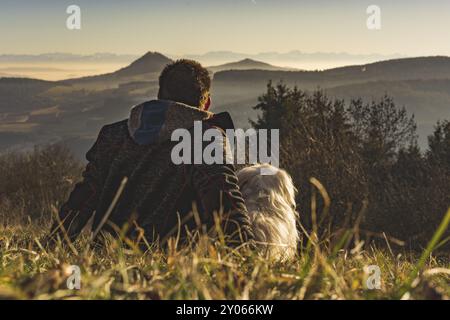 A man sits in the meadow with his dog and looks into the distance Stock Photo