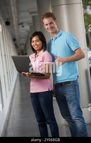 Two happy, smiling college students using a laptop at a gorgeous university campus hallway. 20s female Asian Thai model of Chinese descent. 20s tall m Stock Photo