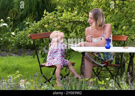 Mother and daughter sitting at a table in the garden. The girl enjoys a tasty snack while her mother looks on in amusement. Relaxed summer atmosphere. Stock Photo