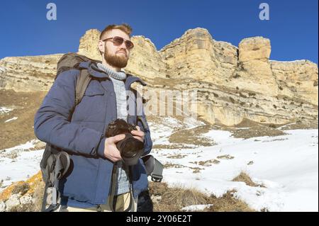 Portrait A bearded hipster photographer with a backpack and wearing sunglasses with a large backpack on his shoulders stands with a DSLR camera in his Stock Photo