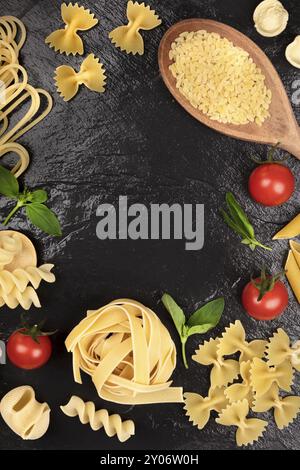 An overhead photo of different types of pasta with basil and cherry tomatoes, shot from above on a black background, forming a frame for copy space Stock Photo