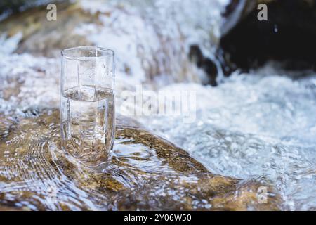 A glass glass with pure mountain drinking water stands on a rock in the course of a mountain river against the backdrop of seething cascades and water Stock Photo