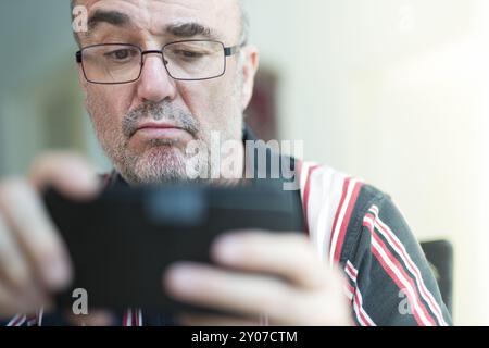 An old man holds a smartphone in his hands and looks sceptically at the display Stock Photo