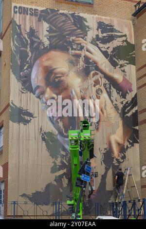 Southend on Sea, UK. 1st Oct, 2024. A large-scale mural in progress on a brick building in Southend-on-Sea, featuring a realistic portrait of a person with braided hair and earrings. Two artists are actively painting—one on a hydraulic lift, the other on a ladder. The mural showcases dramatic lighting and textured background. Artists work to finish their work on the final day of Southend City Jam. Penelope Barritt/Ģ����ý Live News Stock Photo