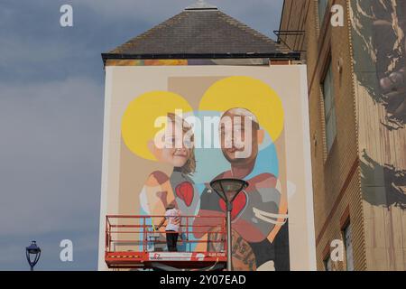 Southend on Sea, UK. 1st Oct, 2024. A large-scale mural being painted on the side of a building in an urban area. The artwork features a child and a tattooed man with stylized hearts and halo-like yellow circles behind their heads. The artist is visible on a red hydraulic lift, actively working on the piece. Artists work to finish their work on the final day of Southend City Jam. Penelope Barritt/Ģ����ý Live News Stock Photo