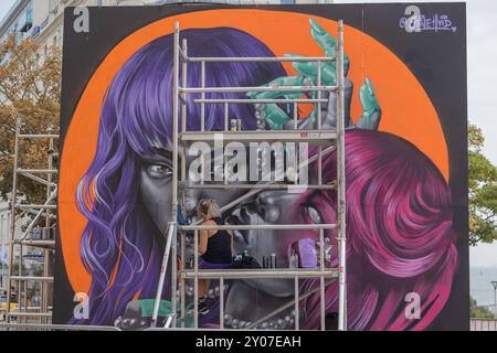 Southend on Sea, UK. 1st Oct, 2024. A street artist works on a large-scale mural featuring two stylized female faces with vibrant purple and magenta hair, surreal green hands, and a bold orange backdrop. The artist is seated on scaffolding, actively painting the wall. Artists work to finish their work on the final day of Southend City Jam. Penelope Barritt/Ģ����ý Live News Stock Photo
