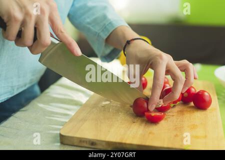 Hands of a young girl chop the cherry tomatoes on a wooden cutting board on a green table in a home setting. The concept of healthy eating and vegetar Stock Photo