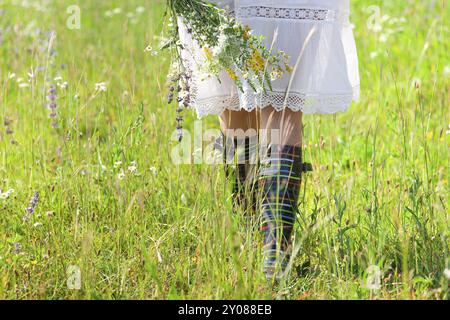 Crop unrecognizable female in rustic white dress and colorful boots with bouquet of wild flowers in hand walking on green field in summer day Stock Photo