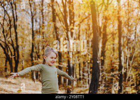 Small happy smiling little caucasian child girl playing with yellow leaves in autumn forest or garden around trees. Kid throws leaves up and they are Stock Photo