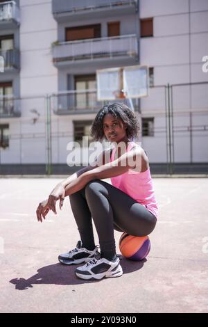 Vertical portrait of an African american woman looking at camera sitting on basketball ball outdoors Stock Photo