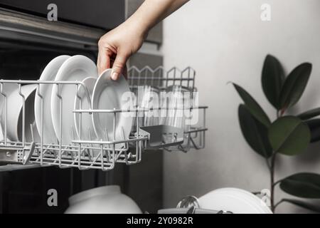 Close-up of female hand loading dished to, empty out or unloading from open automatic stainless built-in dishwasher machine with clean utensils inside Stock Photo