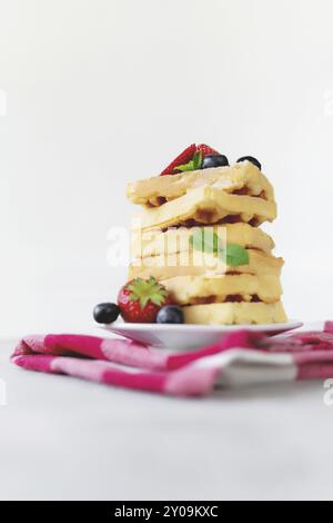 White plate on red kitchen towel with stack homemade Belgian waffles, blueberry, strawberry cuts, mint leaf, sugar powder on gray background Stock Photo