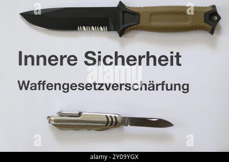 A hunting knife with a beige handle and a Swiss Army knife lie on a white surface next to a text about stricter gun laws Stock Photo