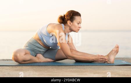 Concentrated young woman making stretching legs sitting on yoga mat and doing exercise yoga on beach at sunrise Stock Photo