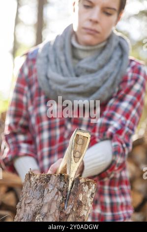 Young woman chopping and splitting logs with an axe on a chopping block. The woman is wearing work or leisure clothes, with woodpiles and forest in th Stock Photo