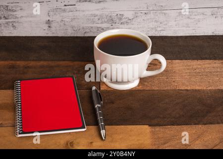 Coffee in white porcelain cup on rustic wood table with red and black covered spiral notebook and ball point pen Stock Photo