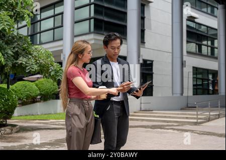 A successful, busy Asian businessman in a formal suit is using a tablet and discussing work with his female assistant while walking outdoors, heading Stock Photo