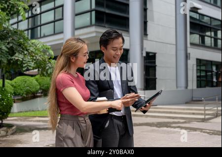 A successful, busy Asian businessman in a formal suit is using a tablet and discussing work with his female assistant while walking outdoors, heading Stock Photo