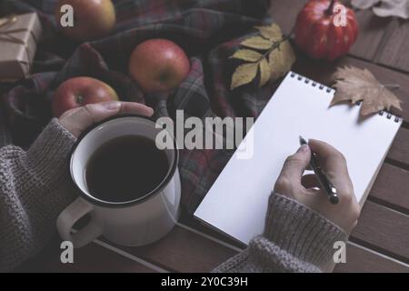 Female hands writing and drinking hot tea checkered blanket apples and autumn leaves on old wooden background Vintage tonning Autumn concept Stock Photo