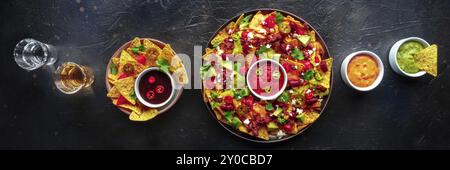 Loaded nachos panorama. Mexican nacho chips with beef, overhead flat lay shot with guacamole sauce, cheese salsa, tequila drinks, on a black slate bac Stock Photo