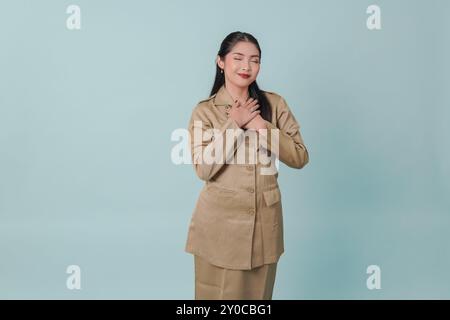 Happy mindful thankful Indonesian government worker woman hand on chest smiling isolated on blue background. PNS civil servant concept. Stock Photo