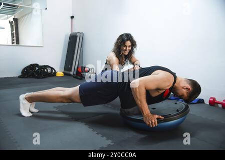 A man doing push-ups on a Bosu ball in a gym with a woman trainer guiding him Stock Photo