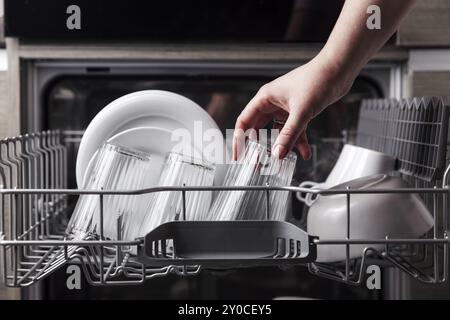 Close-up of female hand loading dished to, empty out or unloading from open automatic stainless built-in dishwasher machine with clean utensils inside Stock Photo
