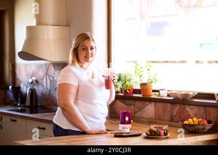 Overweight woman in the kitchen, making fresh smoothie. Choosing healthy food, drinking smoothie from glass. Stock Photo