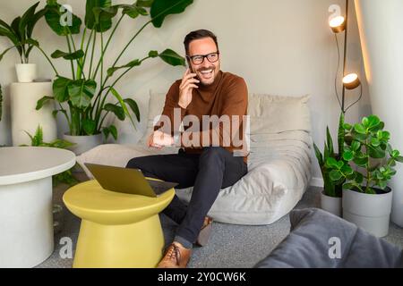 Happy male computer engineer discussing over phone call while working over laptop on sofa in office Stock Photo