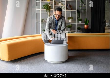 Male project manager in glasses doing online research over laptop while sitting on yellow seat in office Stock Photo