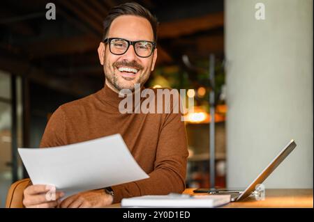 Happy male project manager with document and laptop at desk looking at camera while working in office Stock Photo