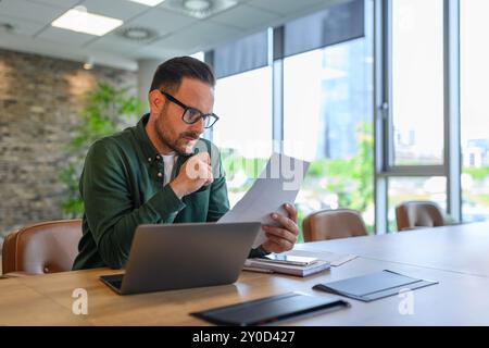 Focused young businessman in glasses reading document while working over computer at desk in office Stock Photo