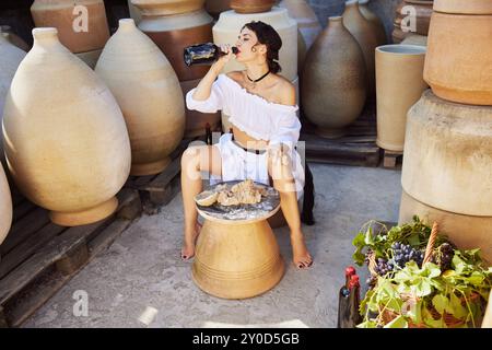 Beautiful young woman working clay in pottery studio, and drinks wine, surrounded by earthy-toned jars. Concept of autumn and harvesting. Stock Photo