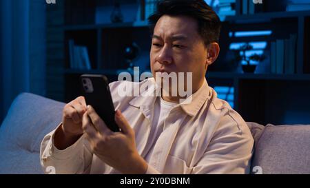 Asian middle-aged man Korean guy mature male evening night home on couch scrolling mobile phone app browsing smartphone chatting social media Stock Photo